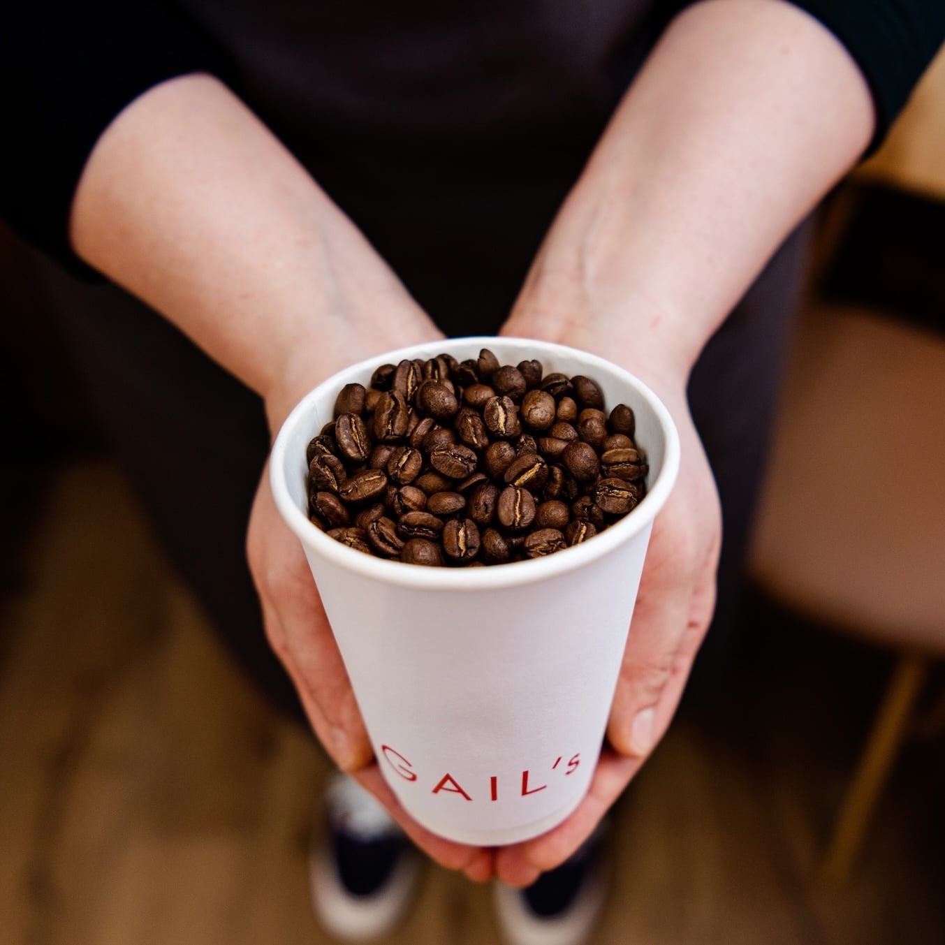 Person holding a cup filled with roasted coffee beans. The cup has "SAIL'S" printed on it.