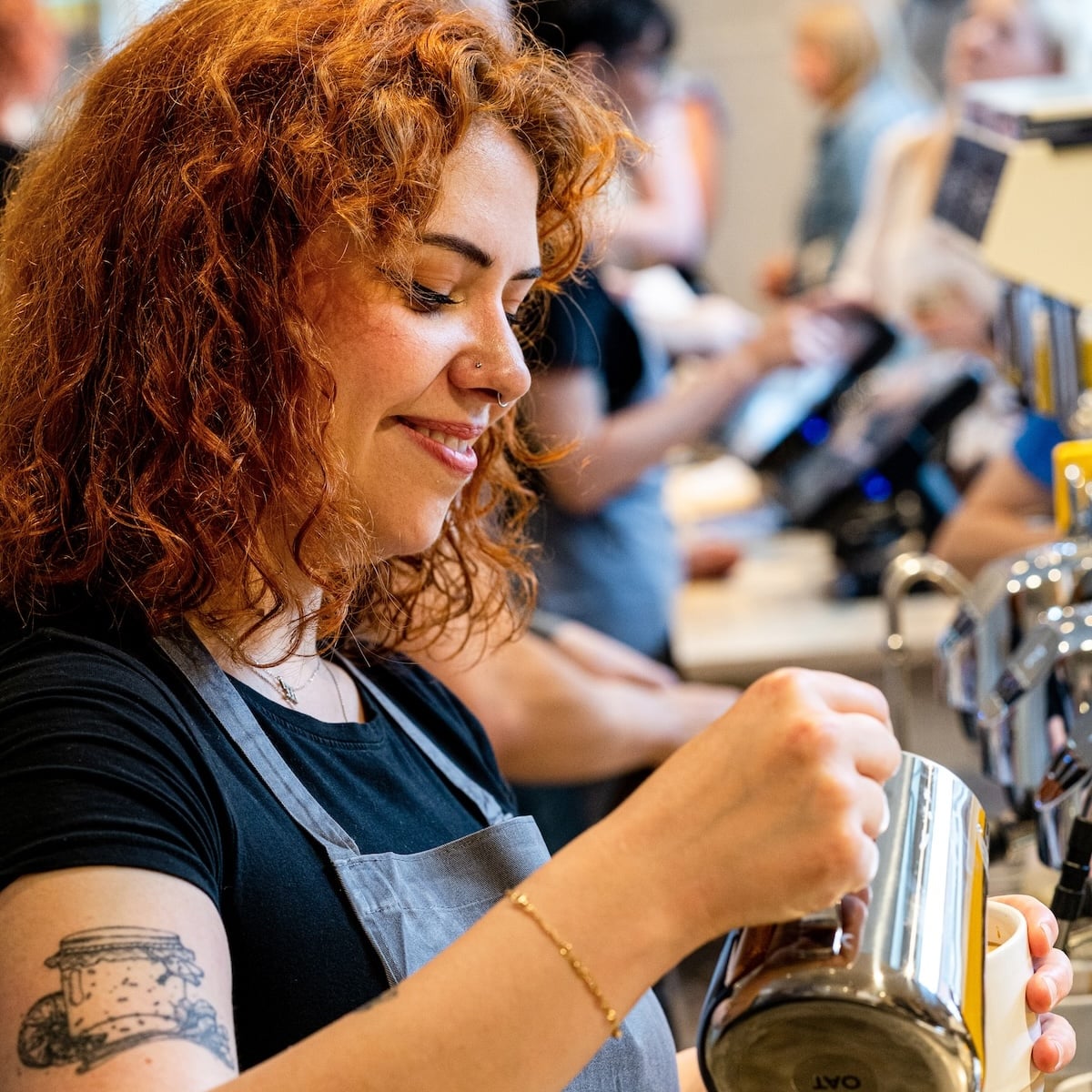 Woman with red hair pouring coffee into a cup. She has a tattoo on her arm.