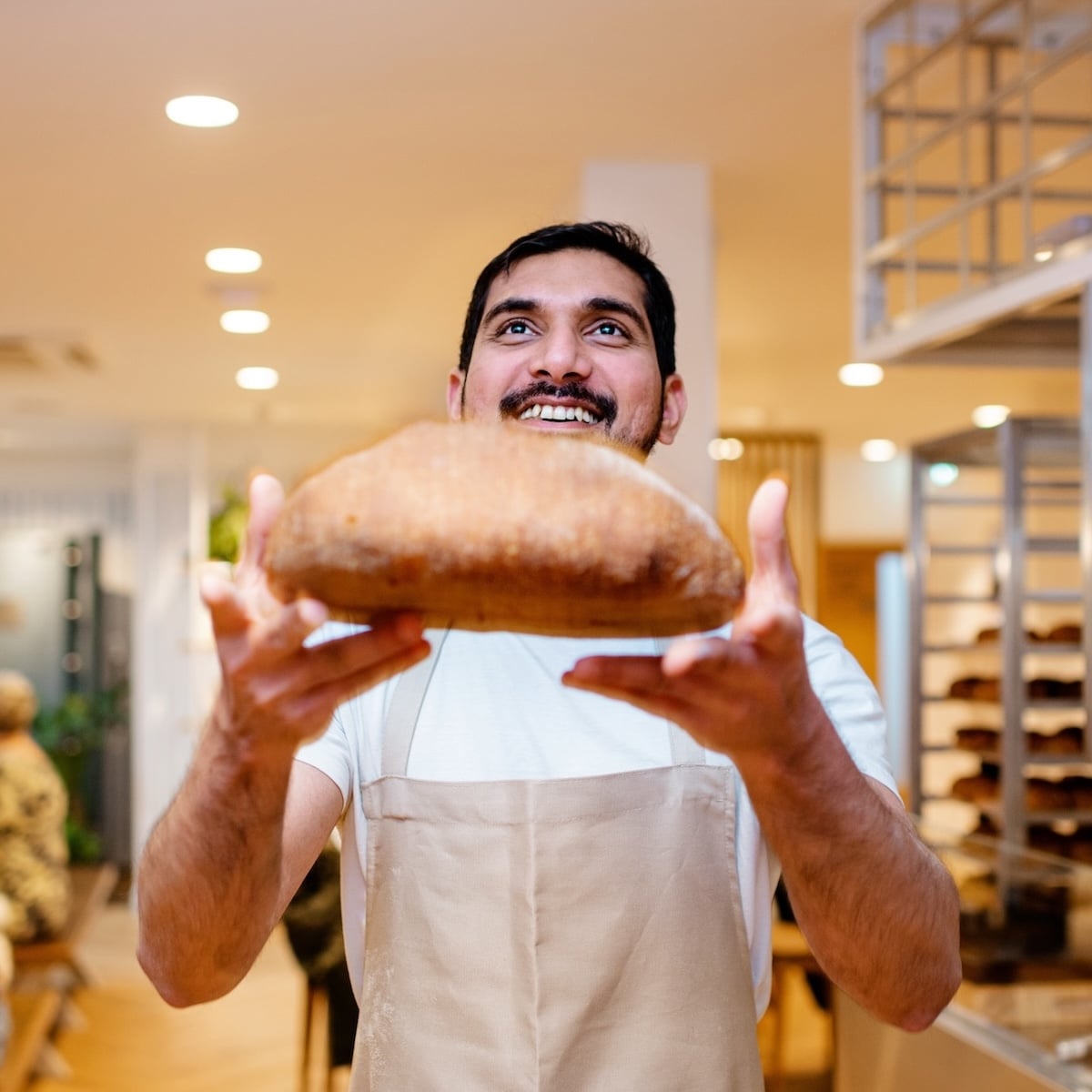 Man holding a loaf of bread, smiling. Wearing an apron.
