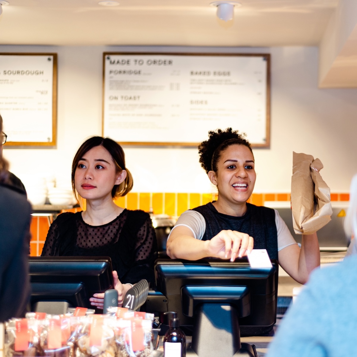 Two women at a cafe counter, one holding a bag, the other looking on.