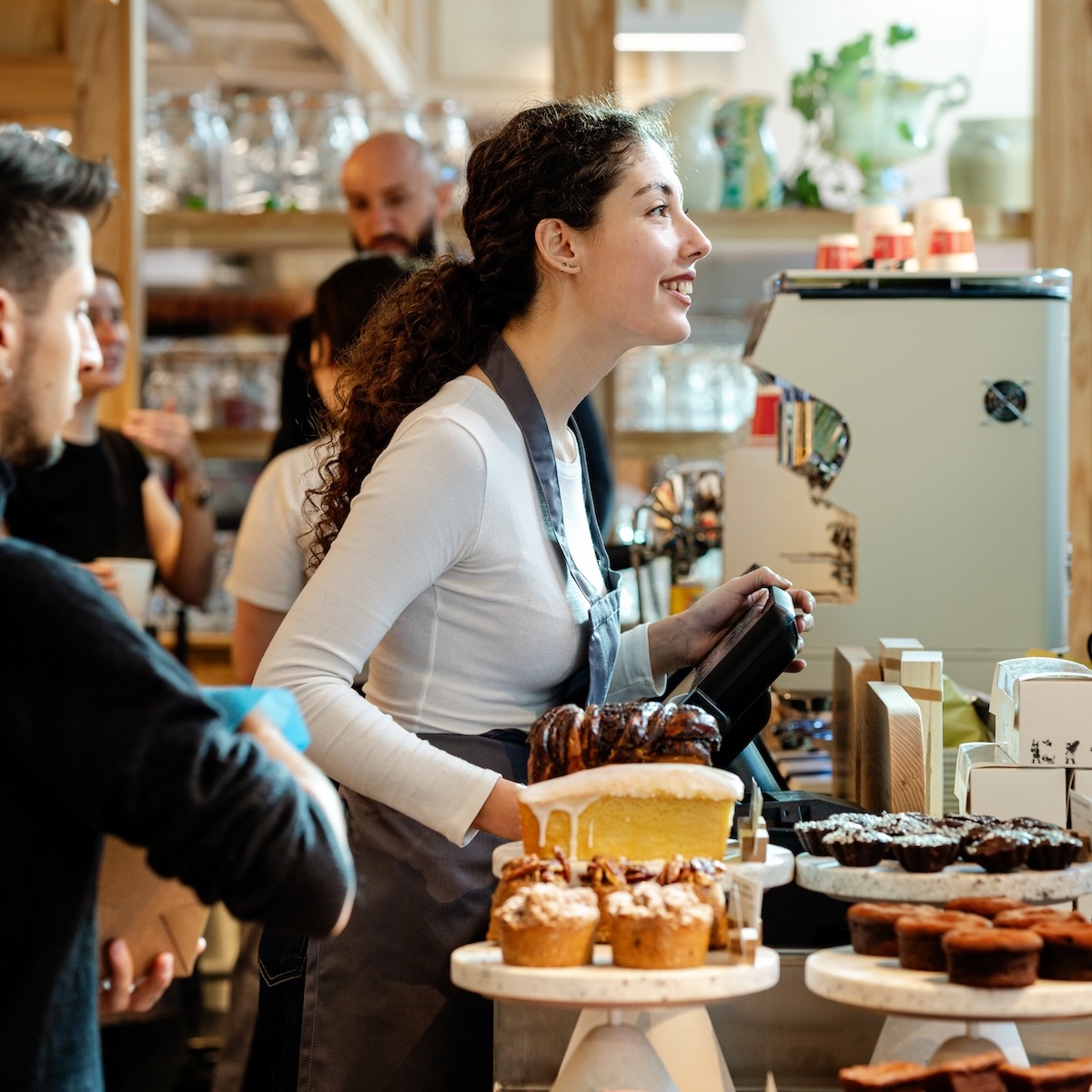 Woman working at a cafe, smiling. Baked goods on display.