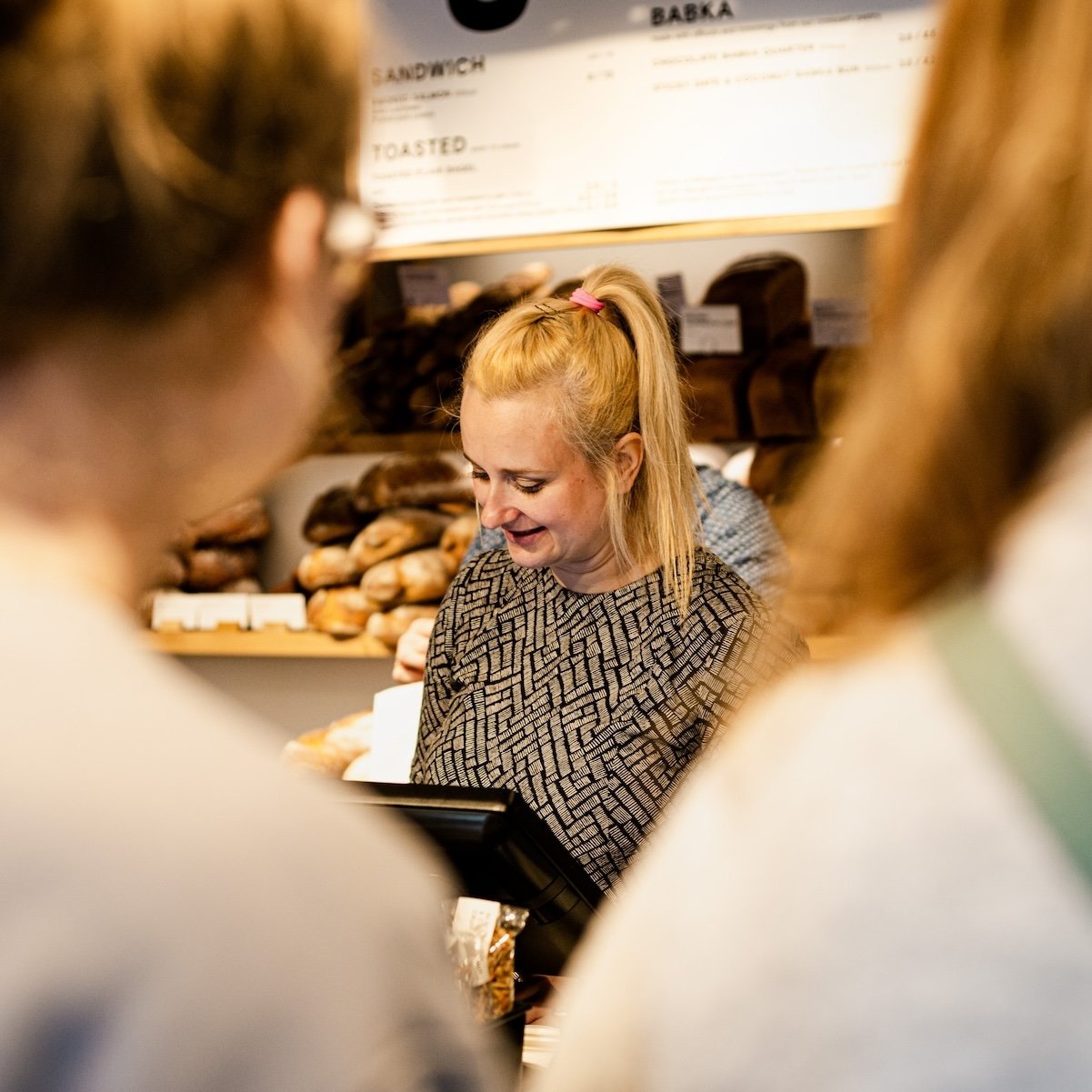 Woman with blonde ponytail at a bakery counter, surrounded by customers.