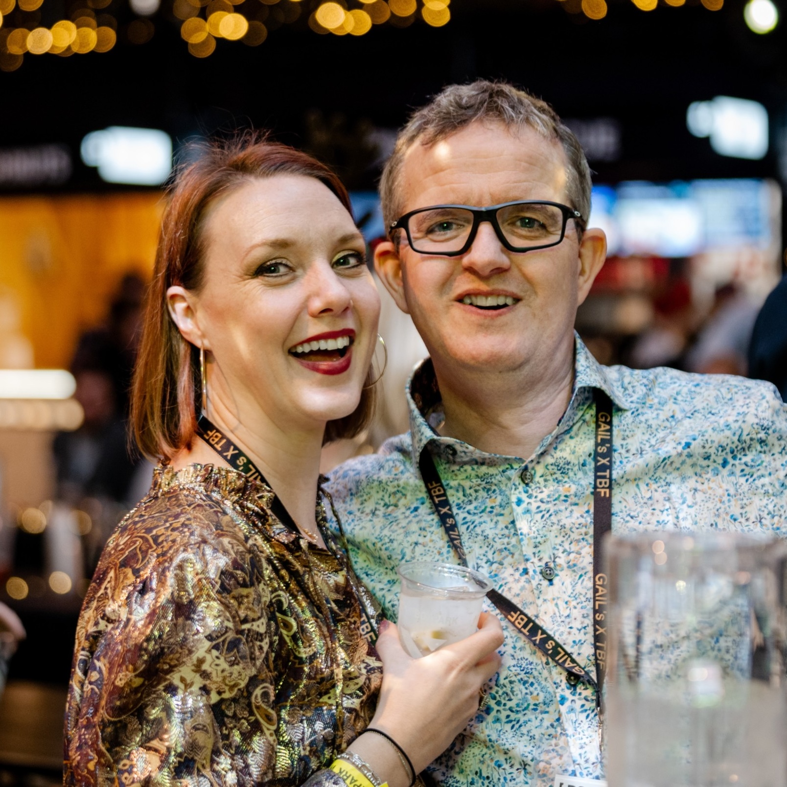 Man and woman smiling, wearing lanyards, holding drinks.