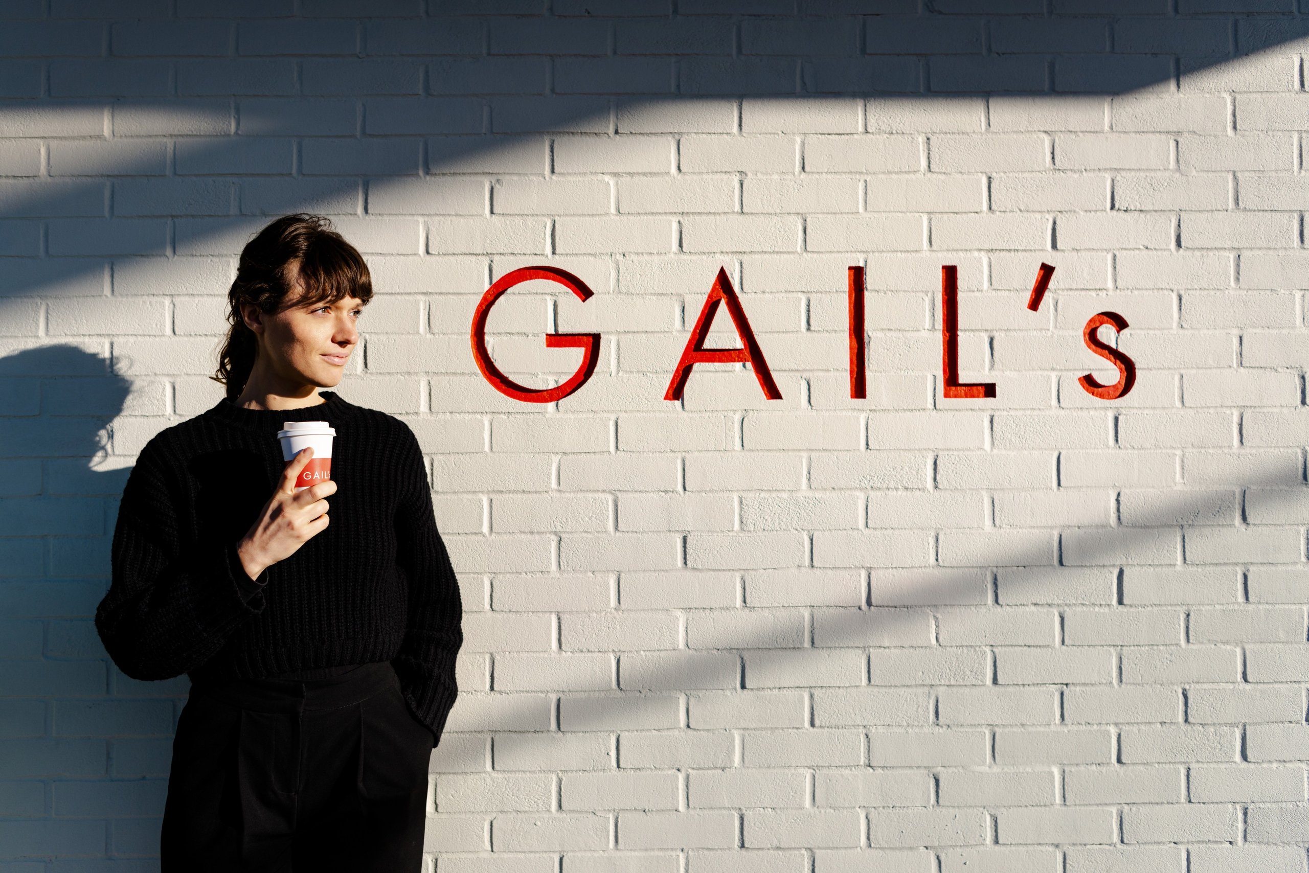 Person holding coffee cup, standing by a brick wall with "Gail's" written on it.