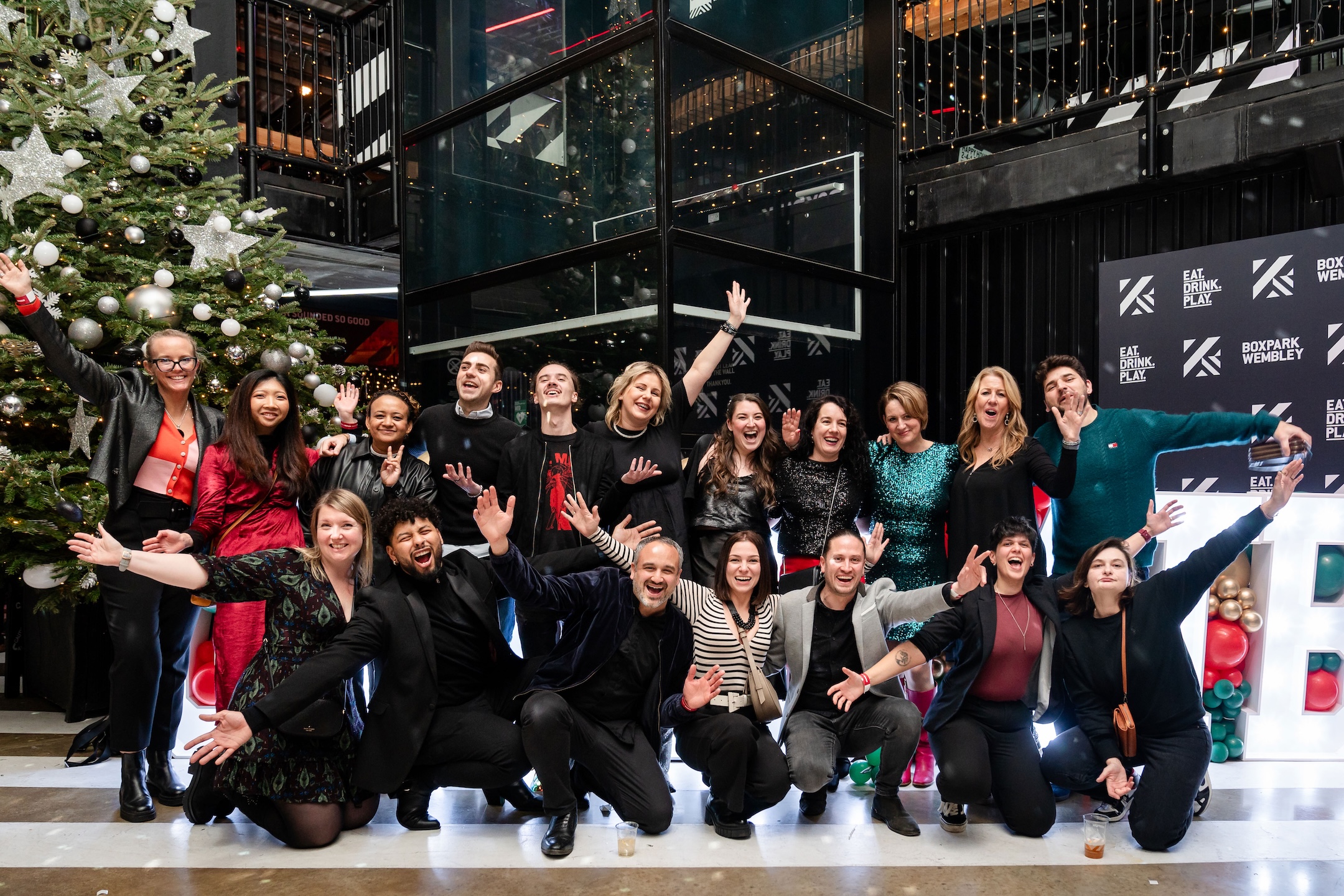 Group of people posing, Christmas tree, "Eat Drink Play" sign.