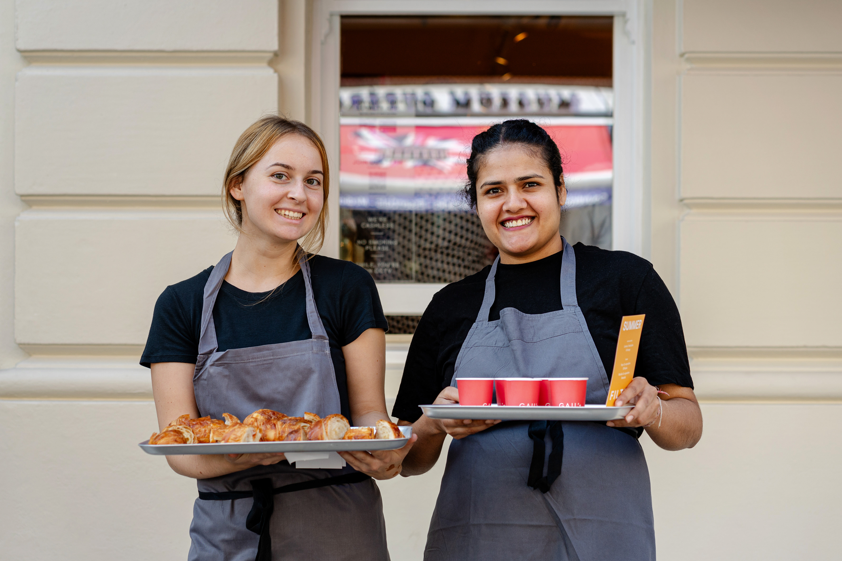 Two women holding trays of food and drinks, smiling at the camera.
