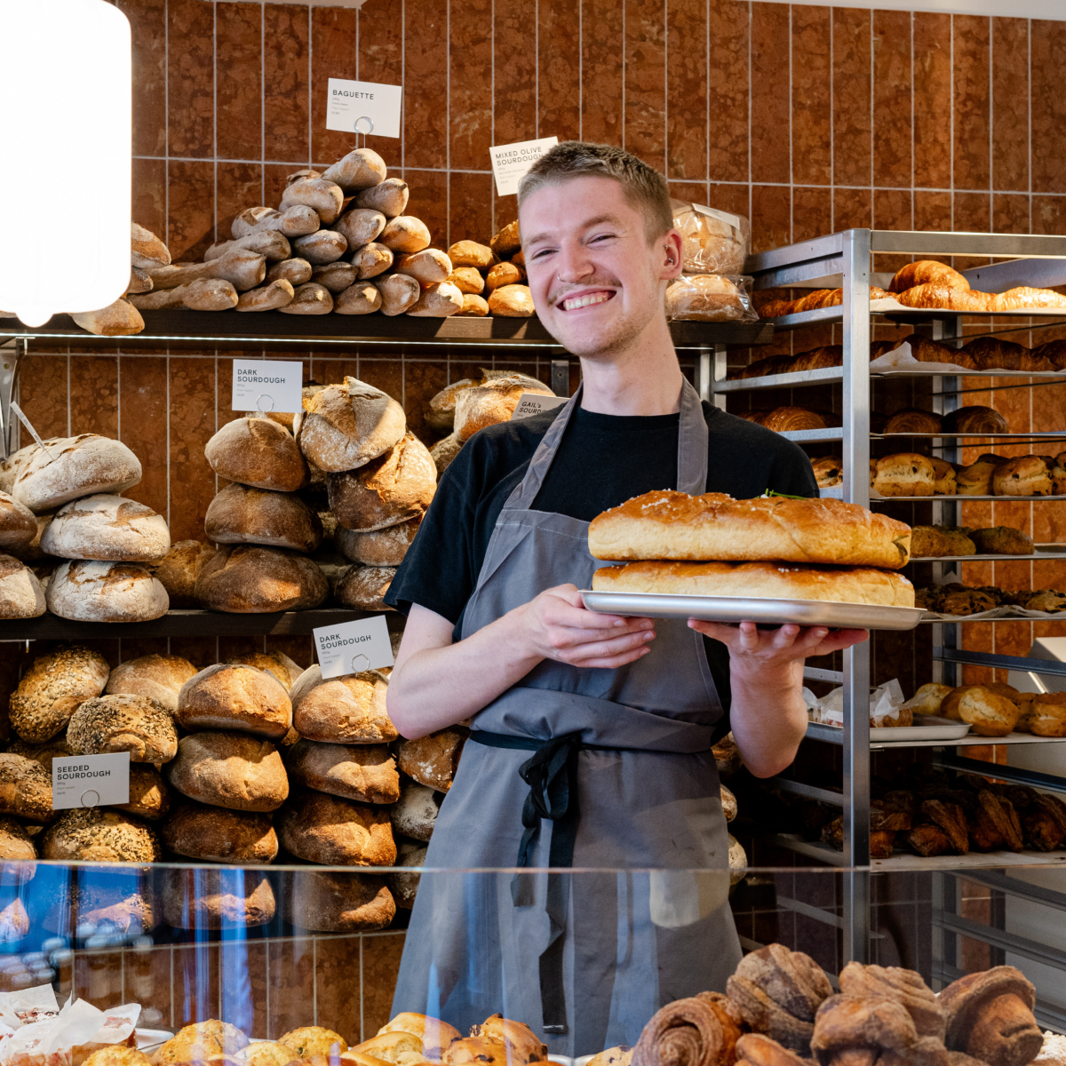 Baker holding tray of bread, smiling in bakery. Shelves of bread in background.