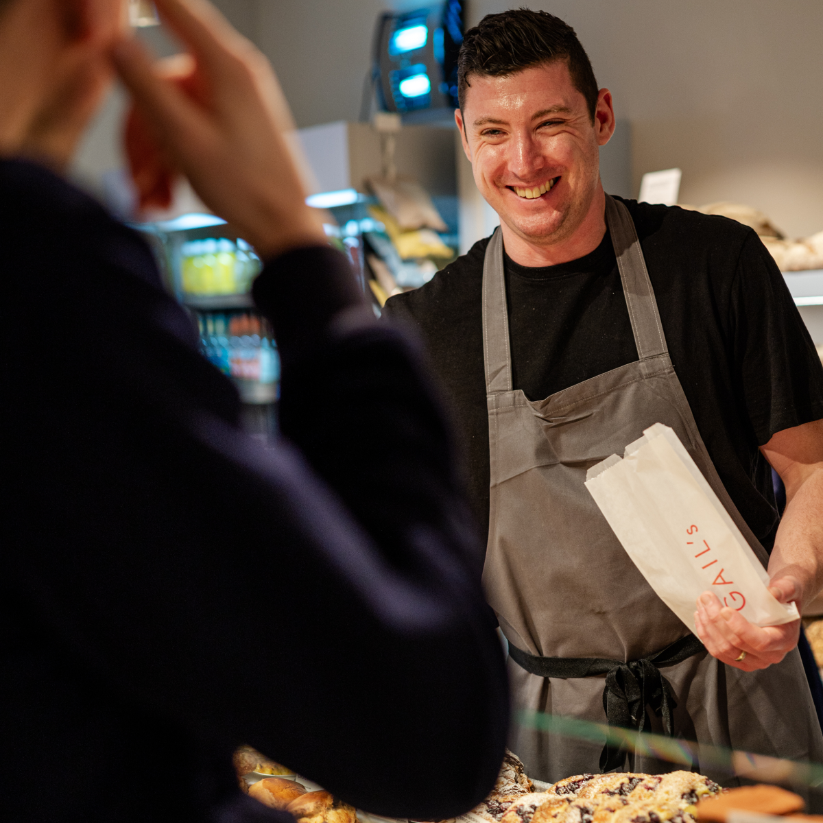 Smiling worker hands a bag to a customer. Bag reads "Gail's".