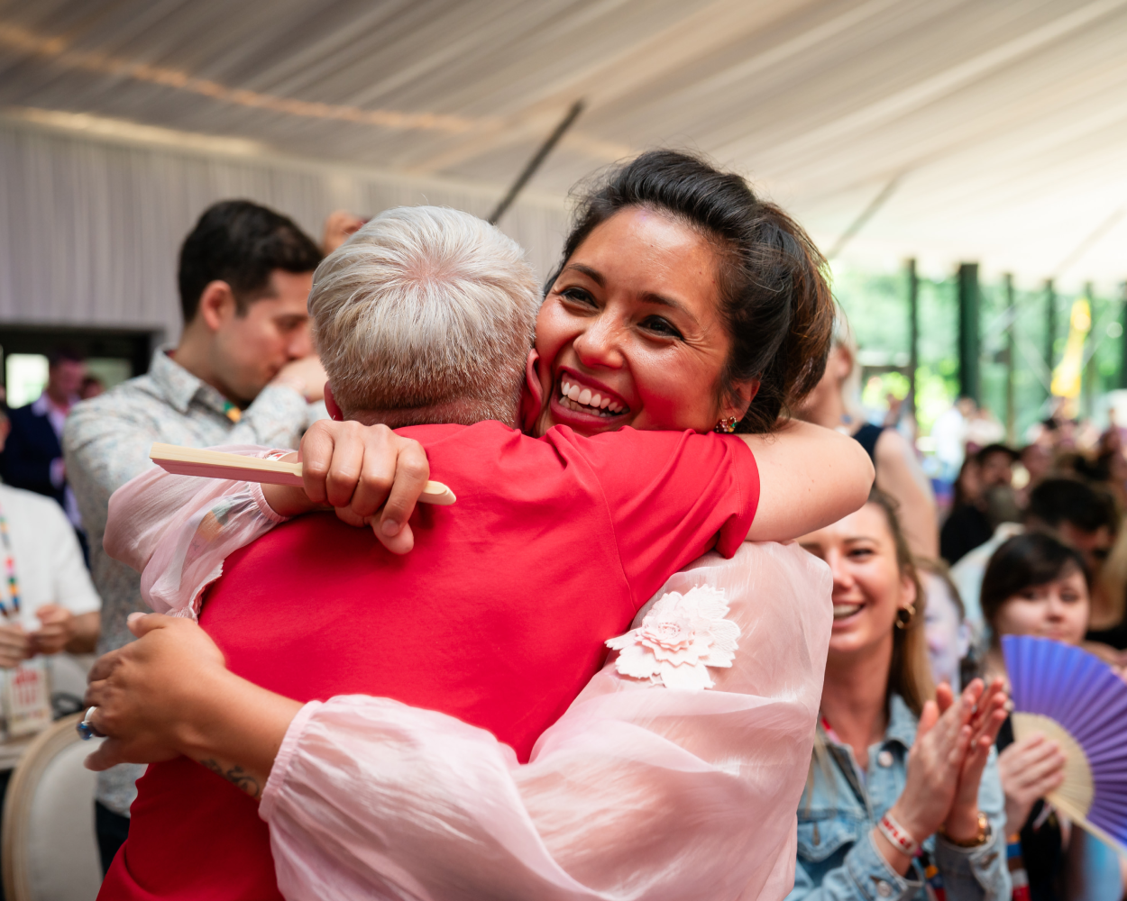 Two women embracing, smiling. People in the background.