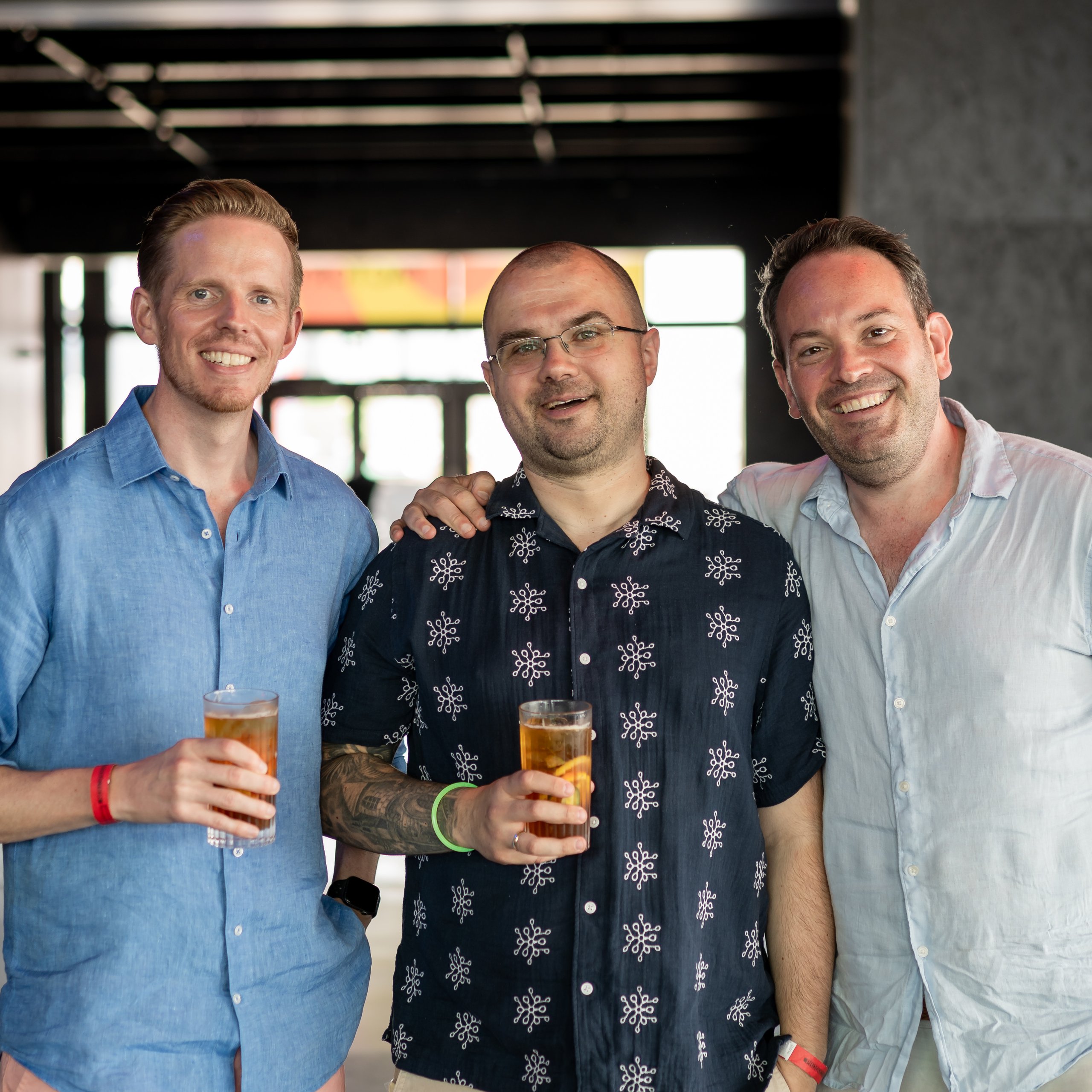 Three men smiling for a photo while holding drinks at an event in a modern, open-plan venue.