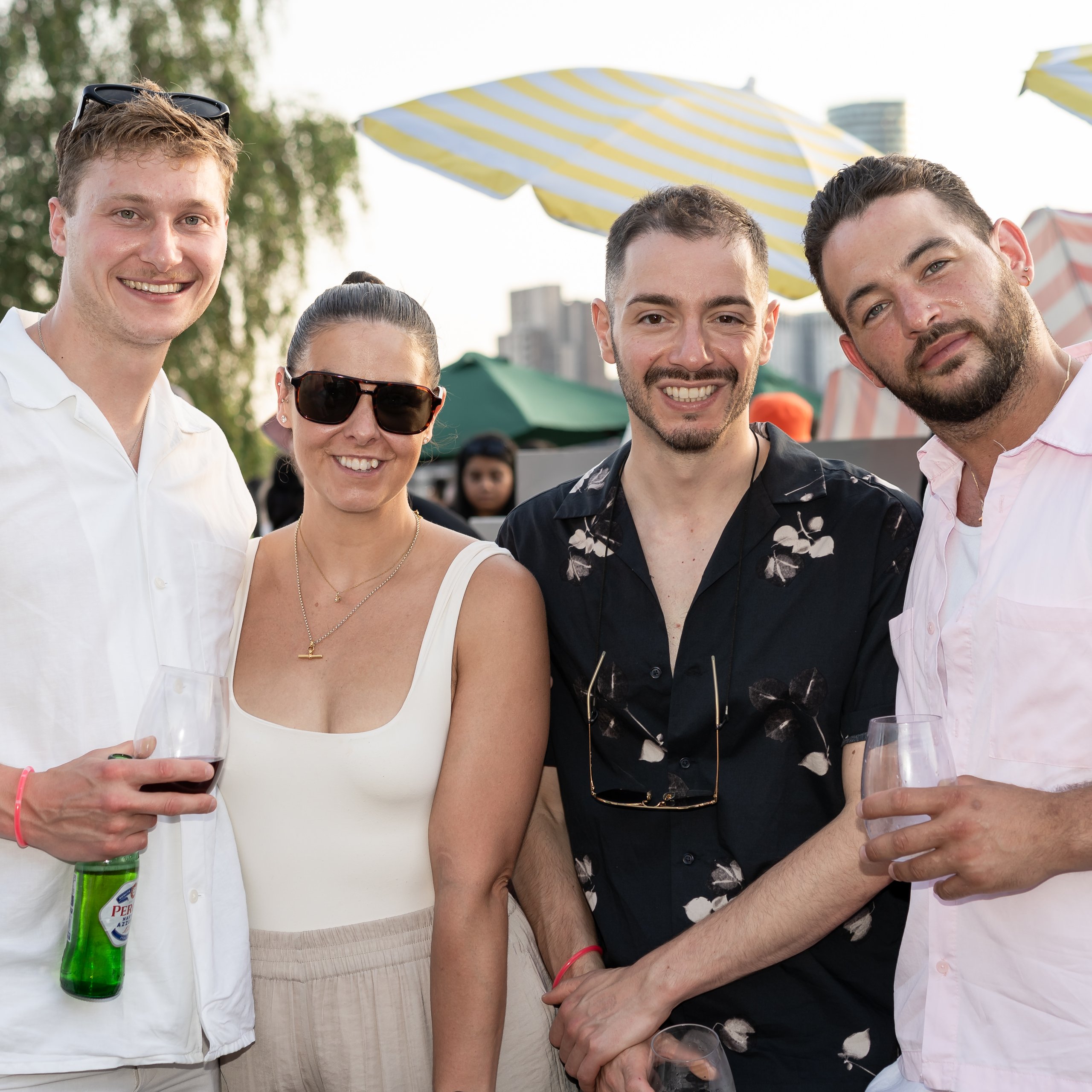 Four friends smiling at an outdoor social gathering with umbrellas in the background.