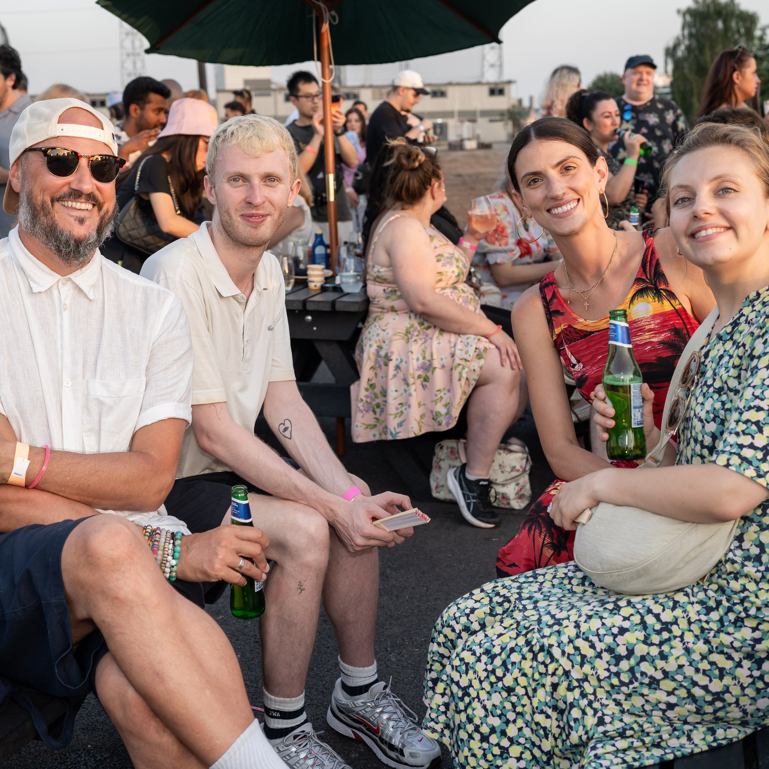 Four friends sitting at an outdoor picnic table with drinks at a sunny gathering.