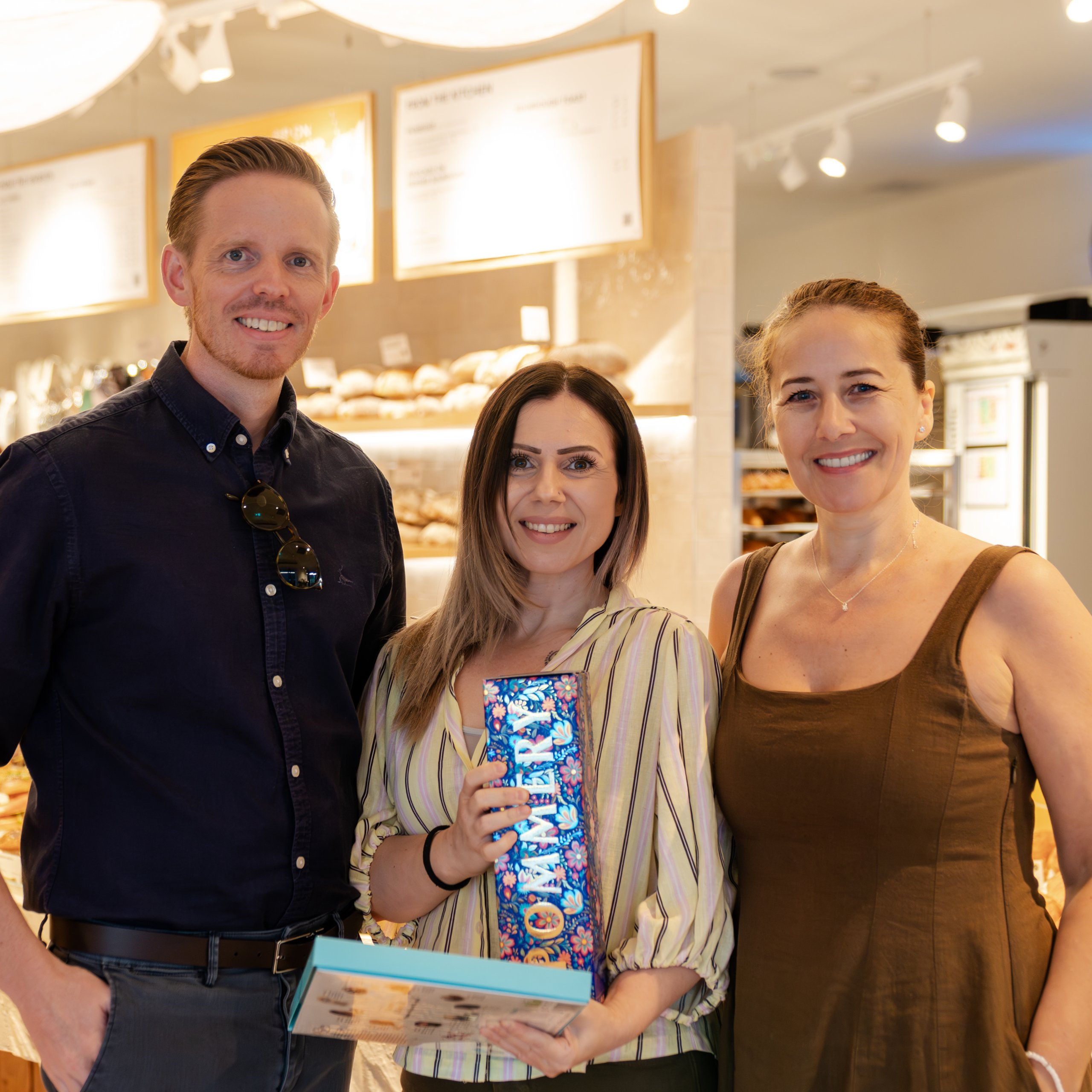 Three people smiling in a bright, modern bakery, one holding a gift box.