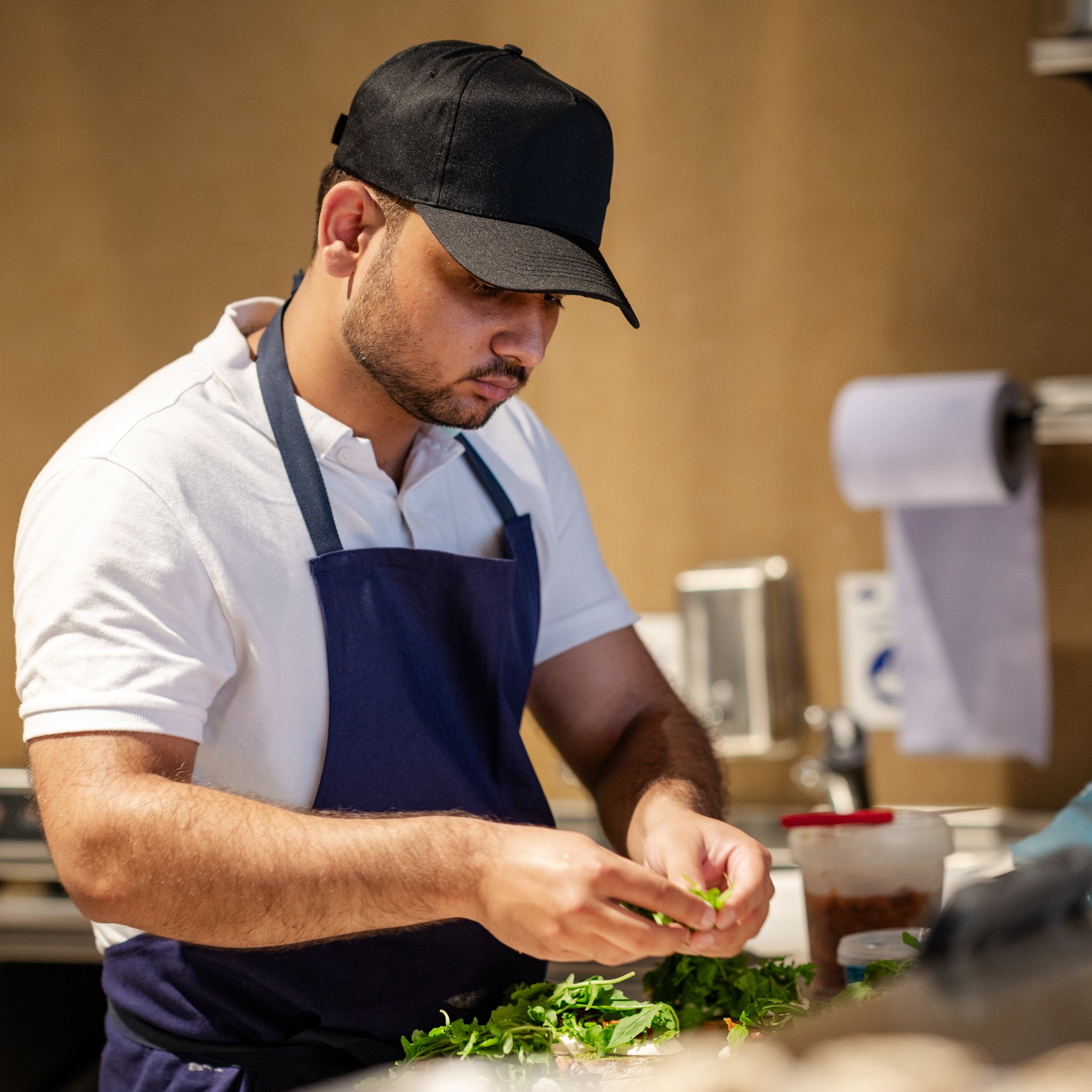 A chef in a white polo and blue apron adds fresh greens to a dish in a professional kitchen.