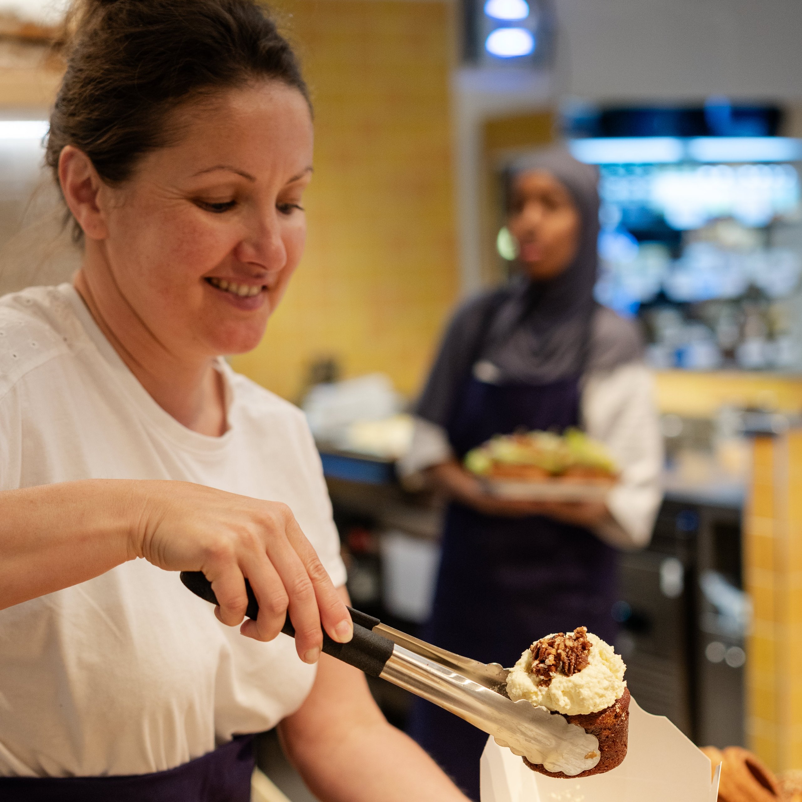 A woman in a bakery uses metal tongs to place a scone with cream and nuts into a takeaway box.