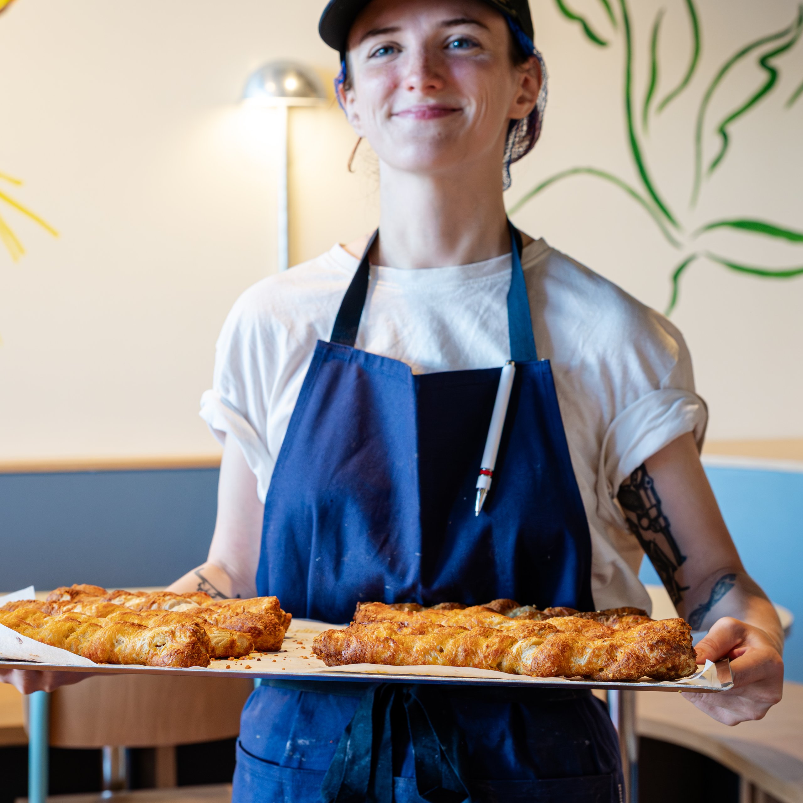A smiling person in a blue apron holds a tray of golden, flaky pastries in a cafe setting.