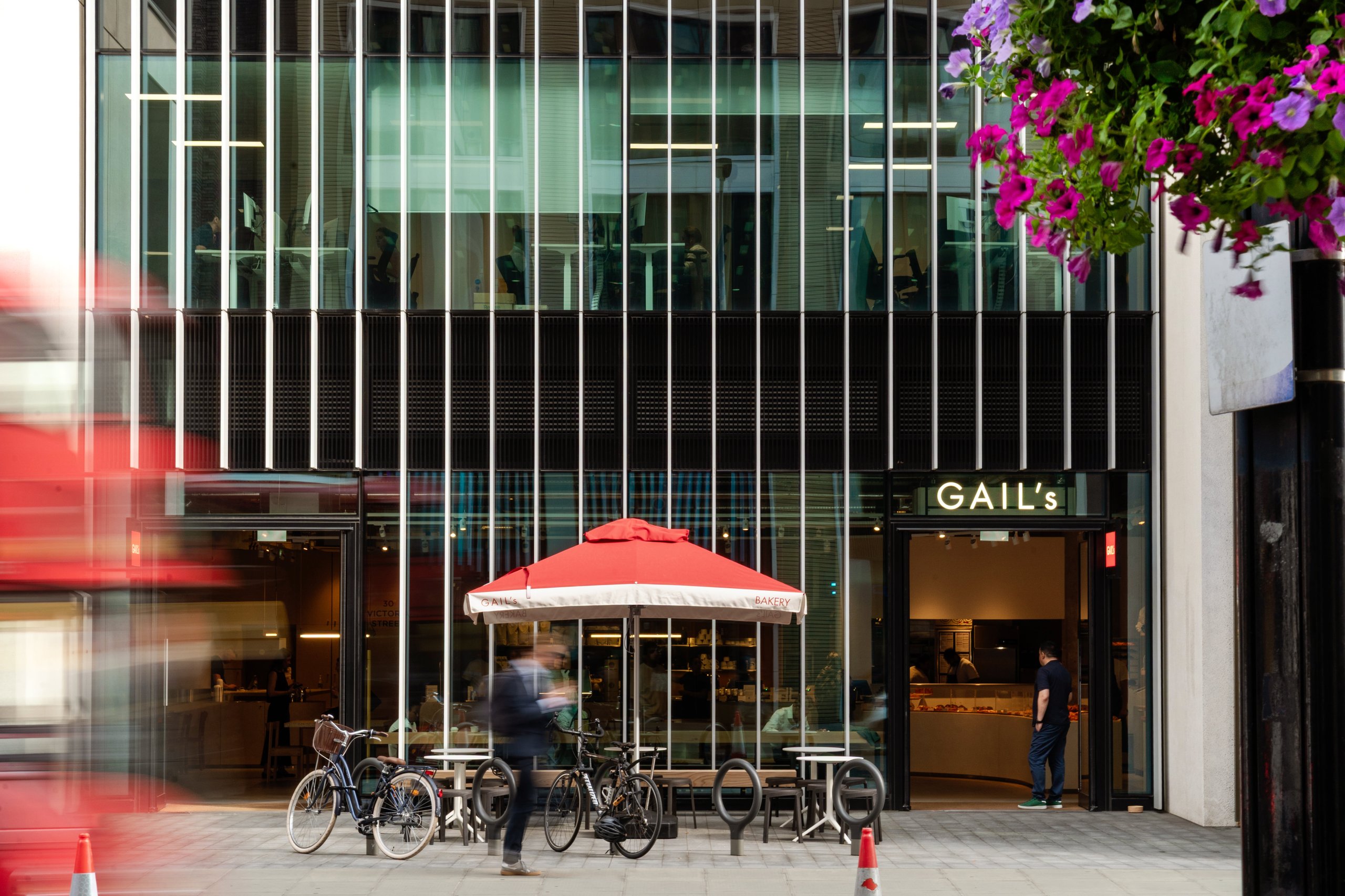 Modern GAIL's Bakery building with a red umbrella, bicycles, and a blurred red bus in London.