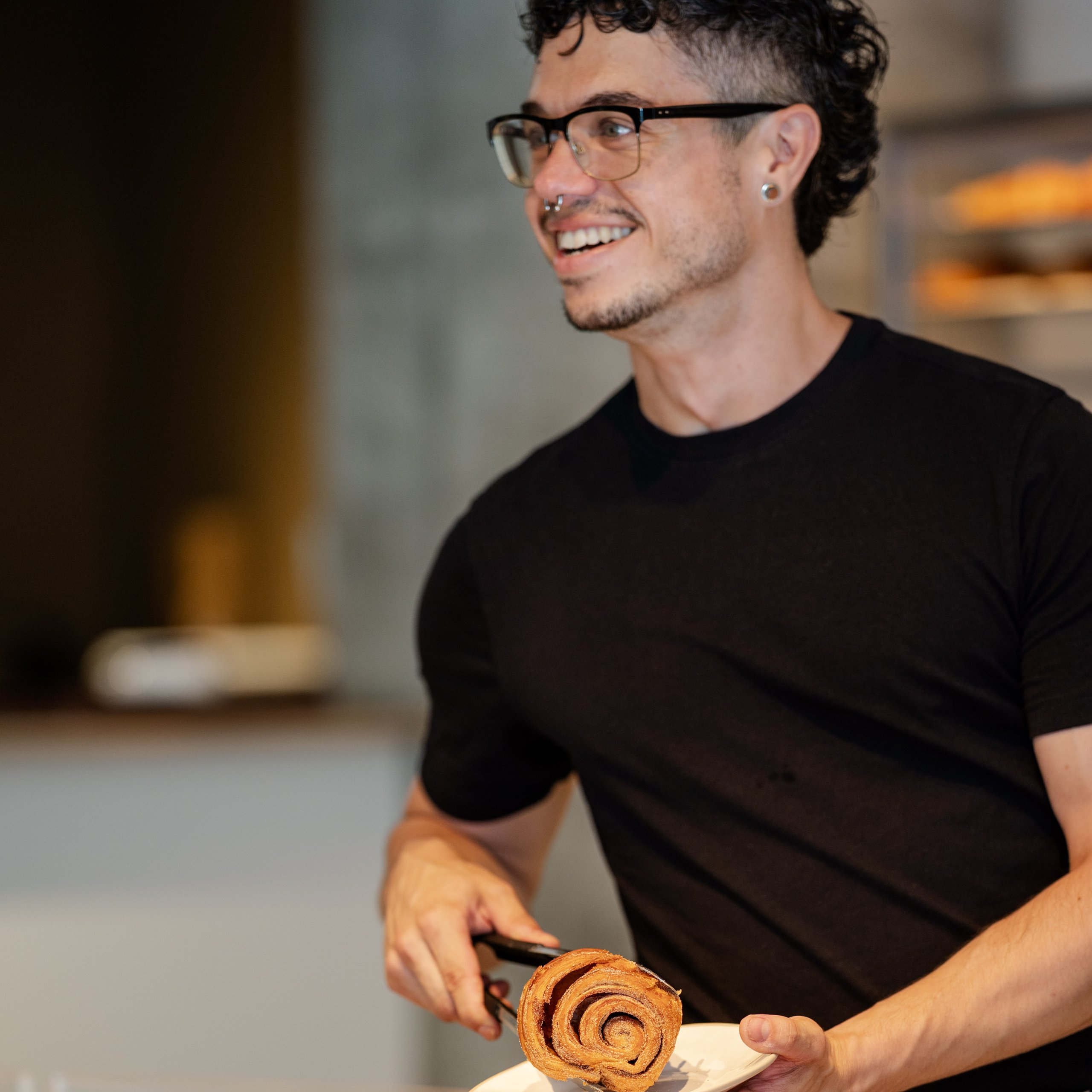 A smiling man wearing glasses and a black t-shirt serves a cinnamon roll on a plate in a bakery.