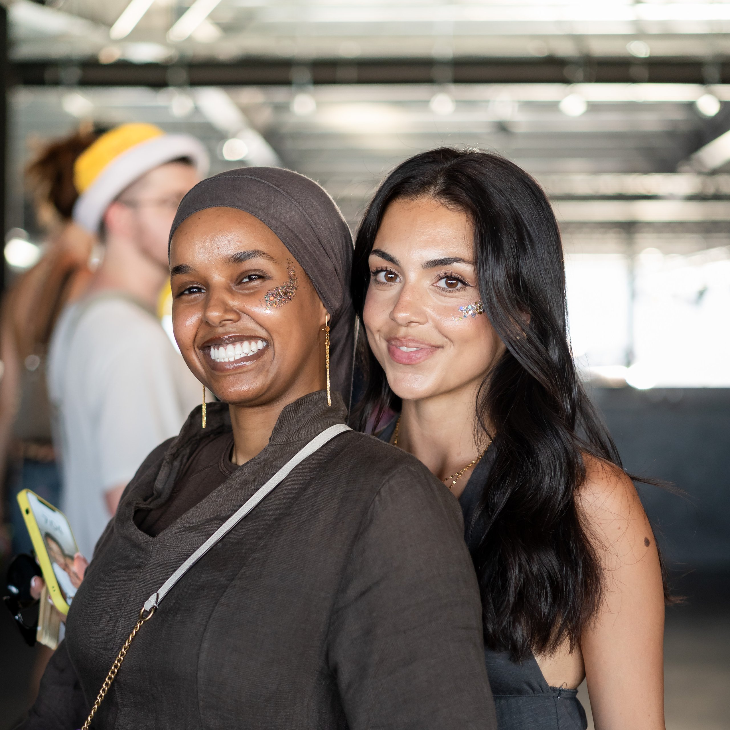 Two young women smiling and posing together indoors, wearing glitter makeup on their cheeks.