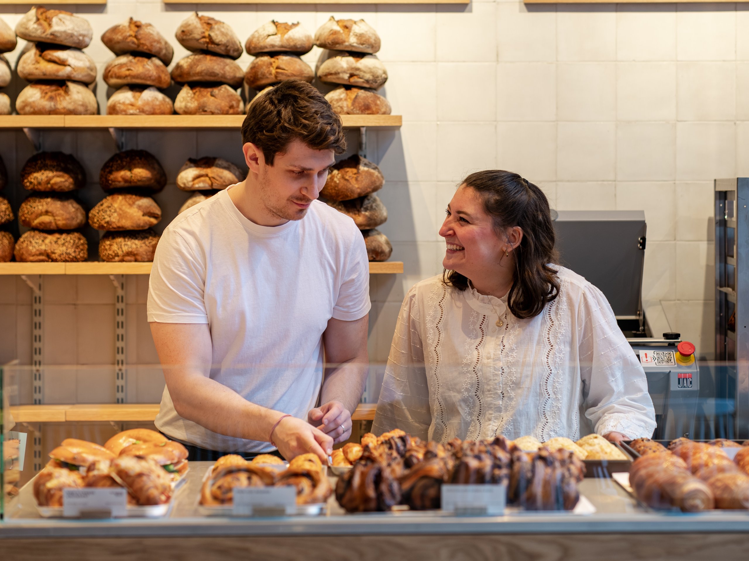 Two smiling bakery workers stand behind a counter filled with pastries, with bread shelves and menus behind them.