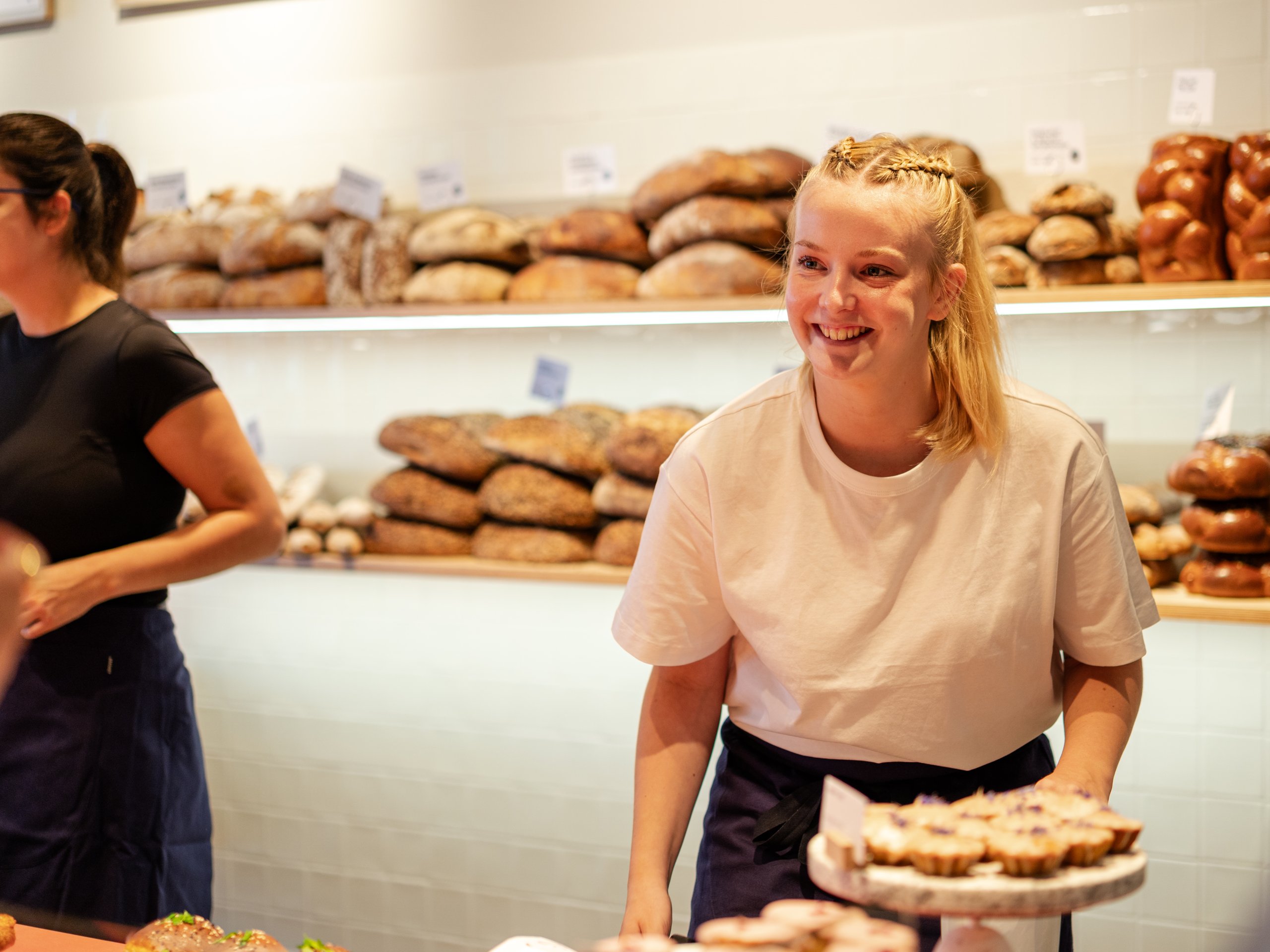 A smiling young female bakery assistant in a white t-shirt holding a tray of pastries.