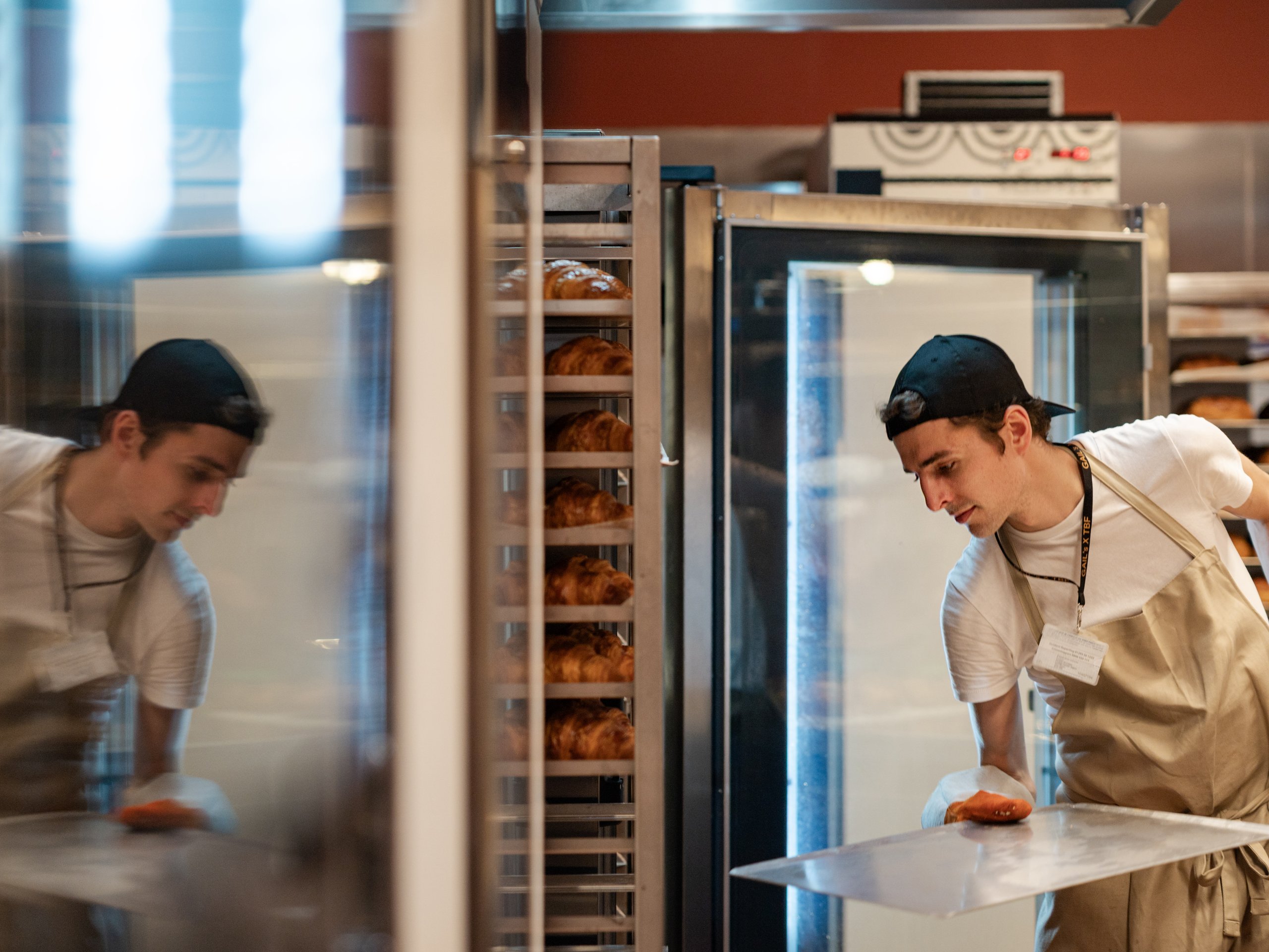 A baker wearing a cap and apron holding a tray near oven shelves filled with baked goods.