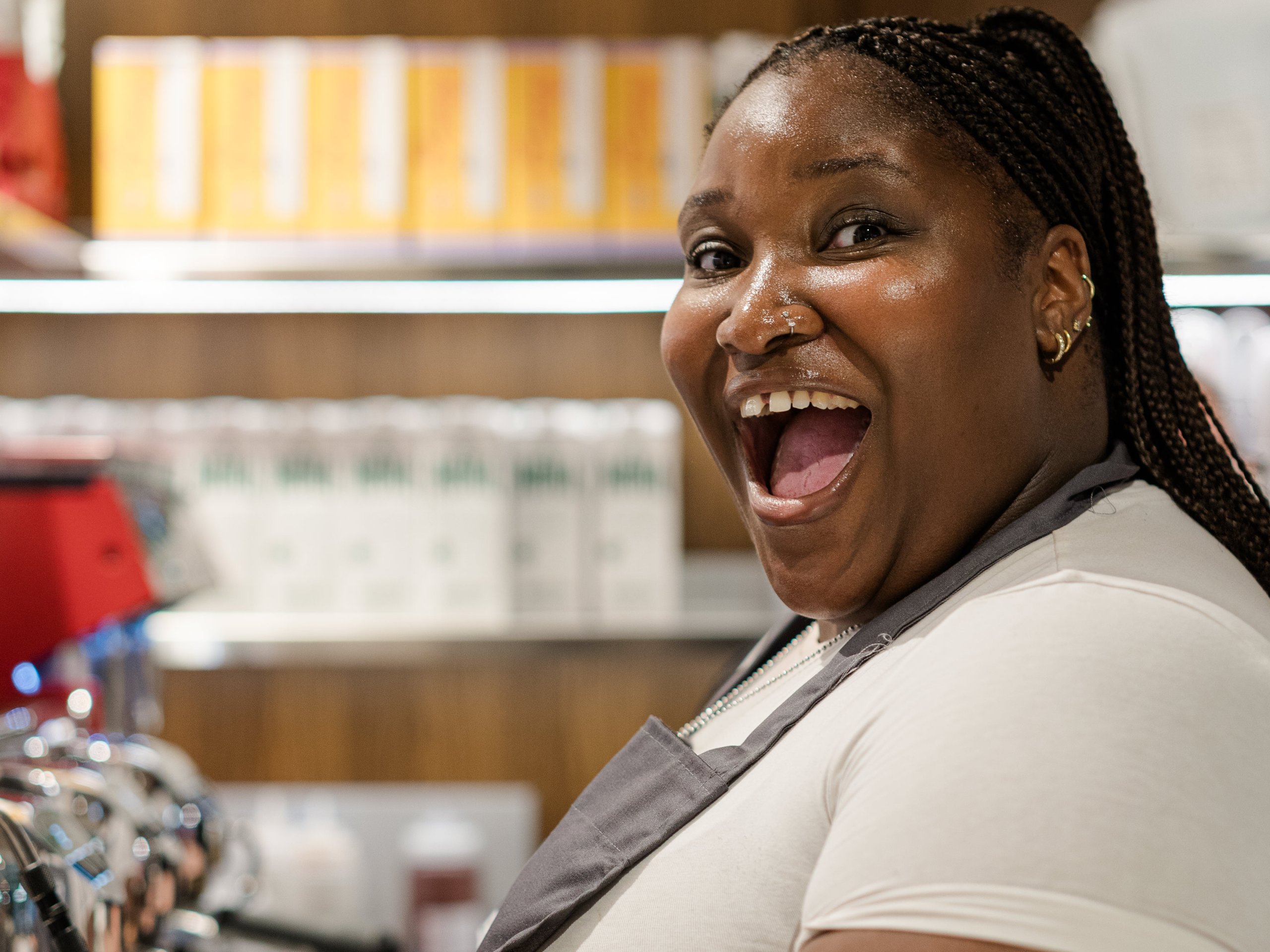A smiling barista with braided hair holding a white cup near an espresso machine in a cafe.