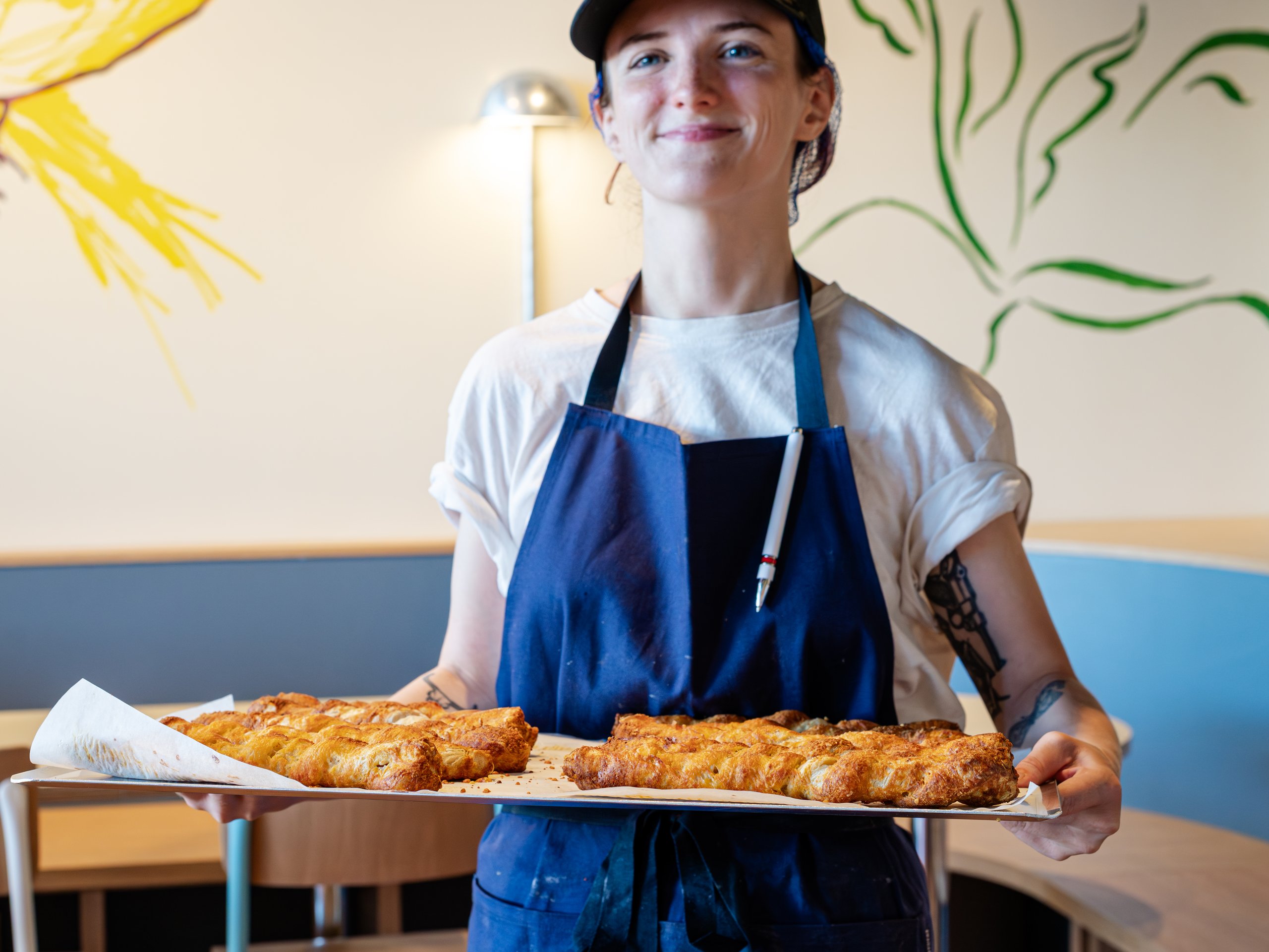 A smiling baker in a blue apron holds a tray of golden brown braided pastries.