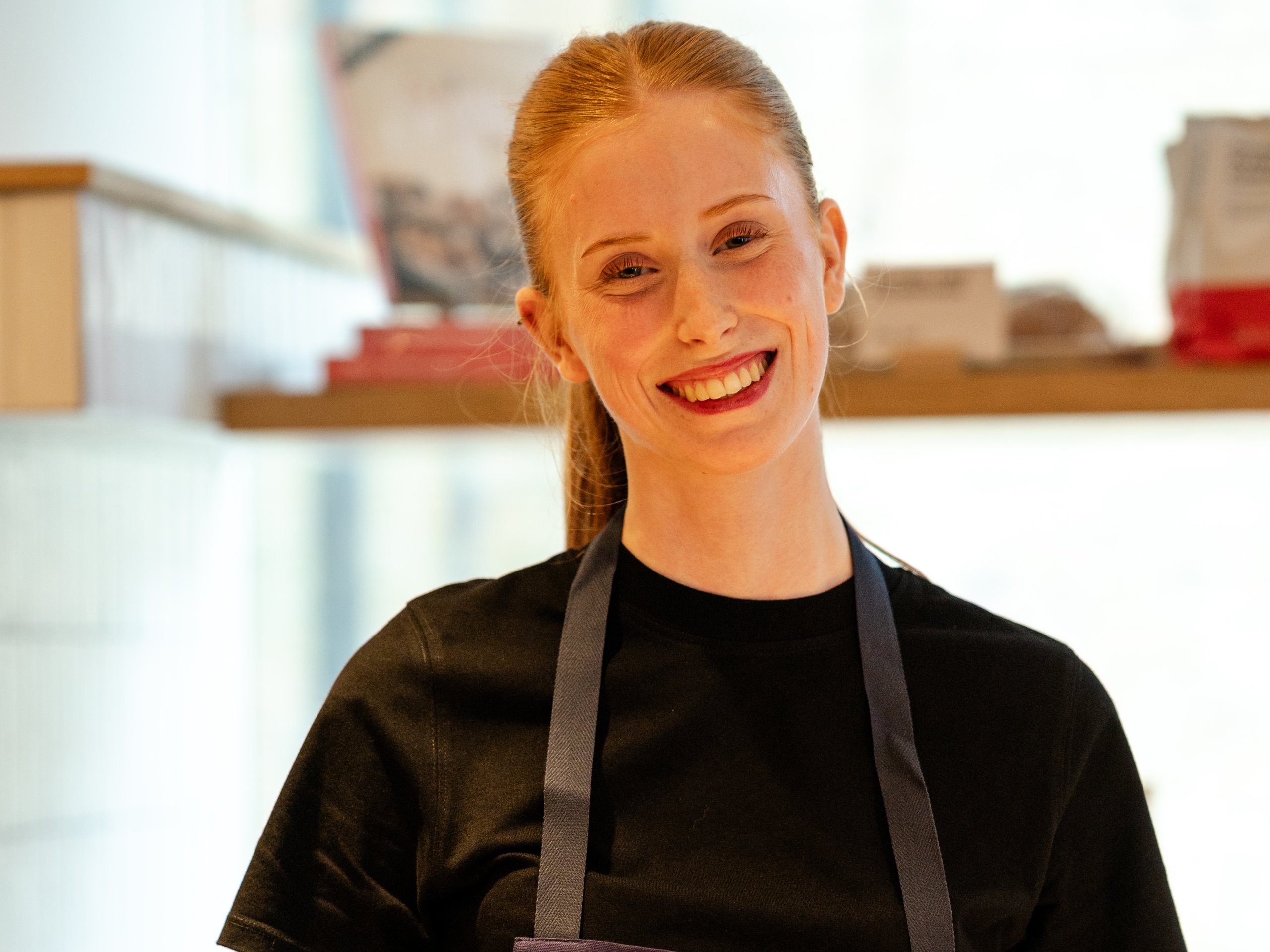 A smiling young female baker with red hair wearing a black shirt and a blue apron in a shop.