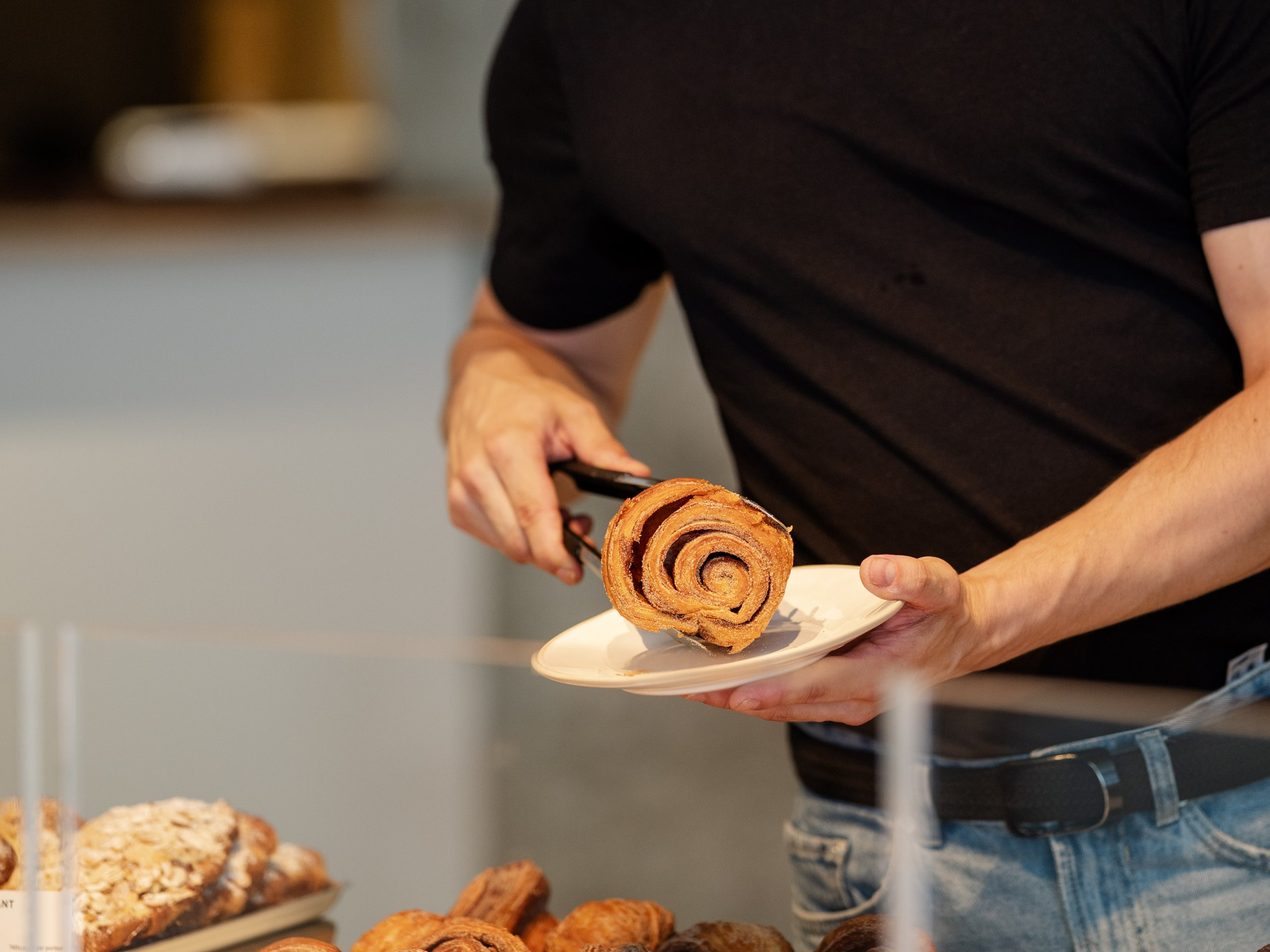A smiling worker in a black t-shirt places a cinnamon bun on a small white plate in a bakery.
