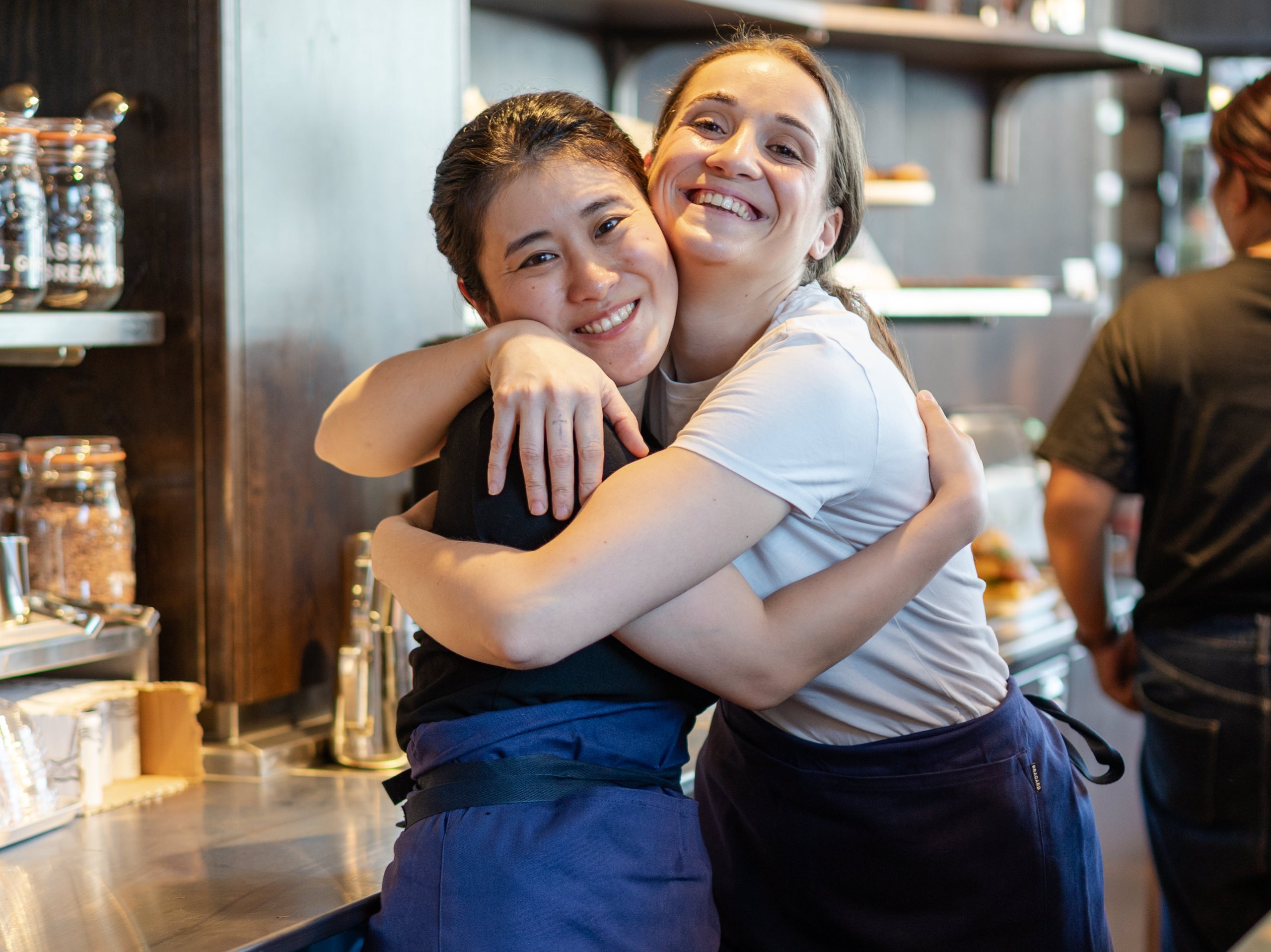 Two female cafe employees with blue aprons joyfully hugging each other behind the counter.