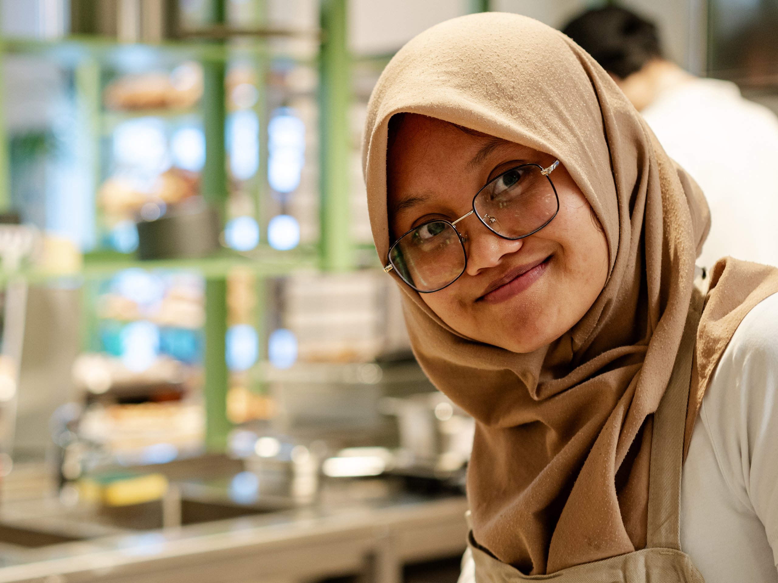 A woman wearing a hijab and glasses smiles while slicing tomatoes and herbs on a cutting board.